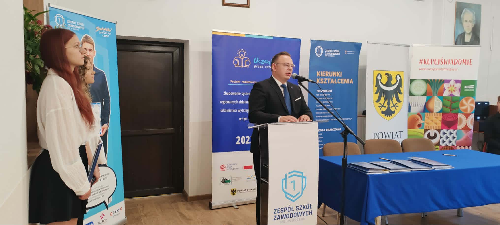 Mężczyzna w garniturze przemawia na mównicy z mikrofonami, obok niebieskiego stołu z dokumentami. Młoda kobieta stoi po lewej stronie banerów. Kolorowe emblematy hrabstwa i szkoły oraz plakaty są widoczne na ścianie za nimi.