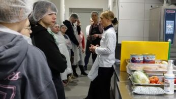 Trzy osoby w foliowych czepkach i fartuchach ochronnych przygotowują jedzenie w profesjonalnej kuchni. Po lewej stronie kobieta kroi marchewkę na zielonej desce, a w centrum mężczyzna w białej czapce kucharskiej pracuje przy żółtej desce. Obok nich stoją puszki z produktami i miska z papryką. Nad głowami widać zawieszone liczne metalowe chochle i przybory kuchenne.