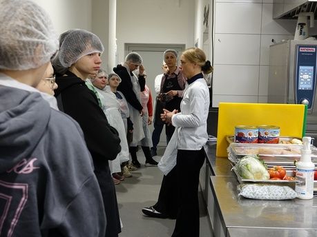 Trzy osoby w foliowych czepkach i fartuchach ochronnych przygotowują jedzenie w profesjonalnej kuchni. Po lewej stronie kobieta kroi marchewkę na zielonej desce, a w centrum mężczyzna w białej czapce kucharskiej pracuje przy żółtej desce. Obok nich stoją puszki z produktami i miska z papryką. Nad głowami widać zawieszone liczne metalowe chochle i przybory kuchenne.