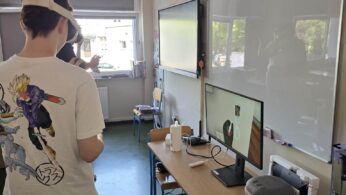Osoba nosząca gogle VR stoi w klasie w pobliżu biurek z komputerem, monitorem, głośnikiem, myszą i kontrolerami VR. Osoba ta ma na sobie białą koszulkę z postaciami z anime. W dobrze oświetlonym pomieszczeniu widoczne są okna, tablica i ekrany.