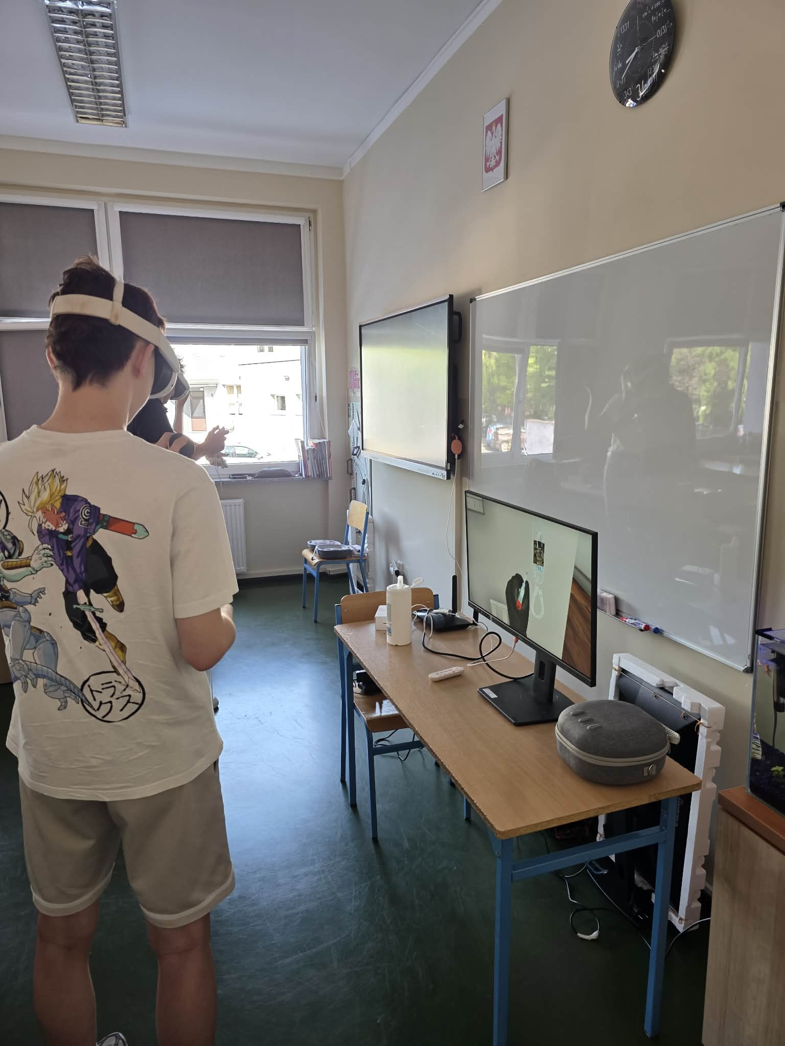 Osoba nosząca gogle VR stoi w klasie w pobliżu biurek z komputerem, monitorem, głośnikiem, myszą i kontrolerami VR. Osoba ta ma na sobie białą koszulkę z postaciami z anime. W dobrze oświetlonym pomieszczeniu widoczne są okna, tablica i ekrany.