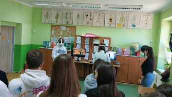 Szerokie ujęcie sali lekcyjnej, w której grupa uczniów obserwuje dwoje rówieśników testujących zestawy VR. Ściany ozdobione są planszami edukacyjnymi przedstawiającymi różne układy ludzkiego ciała. Atmosfera wskazuje na lekcję biologii lub zajęcia techniczne z wykorzystaniem nowoczesnych technologii.