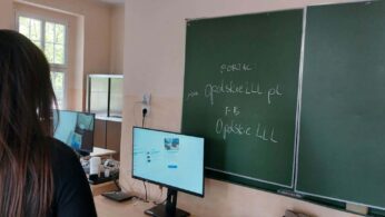 Zdjęcie przedstawia widok zza ramienia osoby patrzącej na monitor komputera oraz zieloną tablicę szkolną. Na tablicy widnieje napis kredą promujący portal 