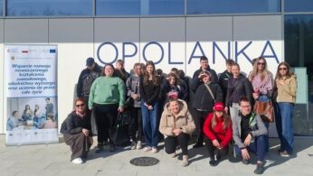 Kobieta w czarnej koszuli i spodniach stoi w centralnej części kadru, gestykulując dłońmi podczas mówienia. Po jej lewej stronie stoi rollup z logotypami i tekstem dotyczącym wspierania kształcenia zawodowego. Za nią, na ekranie projekcyjnym, wyświetlane jest logo z napisem „OPOLANKA RESTAURANT & LEISURE”. Grupa młodzieży siedzi na krzesłach w pierwszym planie, słuchając prezentacji.