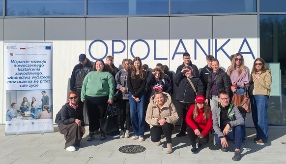 Kobieta w czarnej koszuli i spodniach stoi w centralnej części kadru, gestykulując dłońmi podczas mówienia. Po jej lewej stronie stoi rollup z logotypami i tekstem dotyczącym wspierania kształcenia zawodowego. Za nią, na ekranie projekcyjnym, wyświetlane jest logo z napisem „OPOLANKA RESTAURANT & LEISURE”. Grupa młodzieży siedzi na krzesłach w pierwszym planie, słuchając prezentacji.