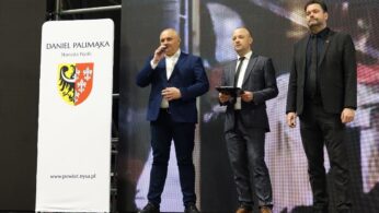 Trzech mężczyzn stoi na scenie przed dużym ekranem LED. Mężczyzna po lewej, w niebieskim garniturze, mówi do mikrofonu. Po prawej stronie sceny stoi biała tablica informacyjna z napisem „DANIEL PALIMĄKA Starosta Nyski”, herbem powiatu i adresem strony internetowej. Scena jest słabo oświetlona, z widoczną metalową konstrukcją w tle.