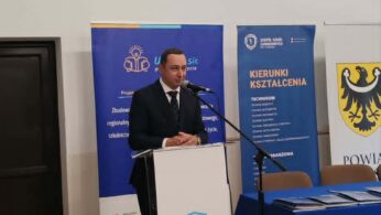 Mężczyzna w garniturze przemawia z podium w pokoju z drewnianą podłogą. Za nim znajdują się niebieskie sztandary i żółty emblemat z czarnym orłem. Stół z niebieskim obrusem i krzesła znajdują się po jego prawej stronie. Nad głową widoczne są światła i zegar.