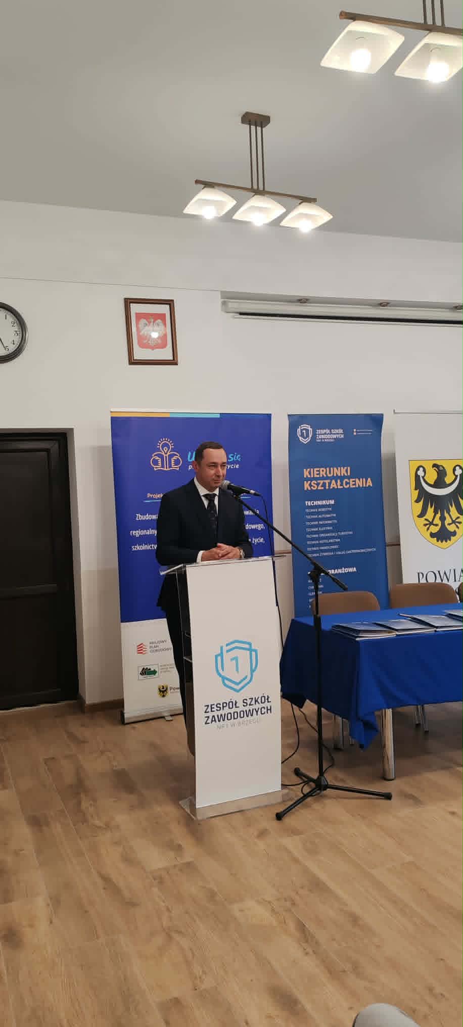 Mężczyzna w garniturze przemawia z podium w pokoju z drewnianą podłogą. Za nim znajdują się niebieskie sztandary i żółty emblemat z czarnym orłem. Stół z niebieskim obrusem i krzesła znajdują się po jego prawej stronie. Nad głową widoczne są światła i zegar.