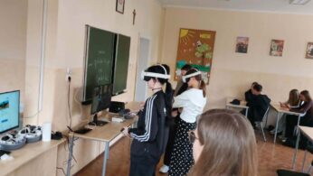 W sali lekcyjnej dwie osoby mają na sobie gogle VR i korzystają z kontrolerów, podczas gdy pozostali uczniowie siedzą przy ławkach w głębi pomieszczenia. Na pierwszym planie widać monitor wyświetlający interfejs portalu internetowego oraz stację dokującą z goglami. Na ścianie obok tablicy wisi gazetka szkolna z napisem 