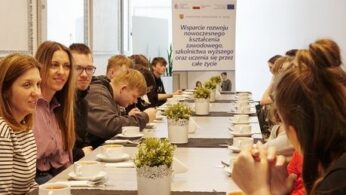 Rząd granatowych toreb płóciennych wisi na oparciach metalowych krzeseł ustawionych w sali. Na każdej torbie znajduje się biały nadruk przedstawiający kopułę i napis „OPOLANKA RESTAURANT & LEISURE”. Pierwsza torba po prawej stronie jest najlepiej widoczna i ostra, pozostałe tworzą linię perspektywiczną w głąb kadru. Po prawej stronie widać fragmenty nóg osób siedzących na krzesłach.
