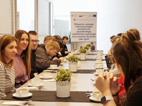 Rząd granatowych toreb płóciennych wisi na oparciach metalowych krzeseł ustawionych w sali. Na każdej torbie znajduje się biały nadruk przedstawiający kopułę i napis „OPOLANKA RESTAURANT & LEISURE”. Pierwsza torba po prawej stronie jest najlepiej widoczna i ostra, pozostałe tworzą linię perspektywiczną w głąb kadru. Po prawej stronie widać fragmenty nóg osób siedzących na krzesłach.