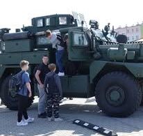 Czworo nastolatków w swobodnym ubraniu ogląda duży, wojskowy pojazd opancerzony zaparkowany na zewnątrz na brukowanym placu. Jeden chłopak wspina się po stopniach do otwartych drzwi pojazdu, a pozostali obserwują. Pojazd jest ciemnozielony, z dużymi oponami i pancernymi oknami. W tle widać niewyraźne budynki mieszkalne.