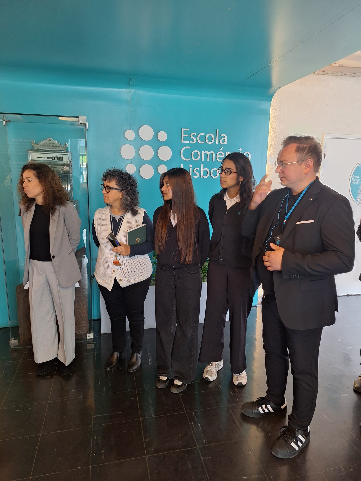 Pięć osób stoi w rzędzie w nowoczesnym wnętrzu na tle jasnoniebieskiej ściany z białym napisem „Escola Comércio Lisboa”. Wśród nich znajdują się trzy kobiety i dwóch mężczyzn, z których jeden po prawej stronie gestykuluje dłonią podczas wypowiedzi. Po lewej stronie, za szybą, widoczna jest zabytkowa, metalowa kasa fiskalna. Cała grupa jest ubrana w eleganckie, casualowe stroje w ciemnych i szarych kolorach.