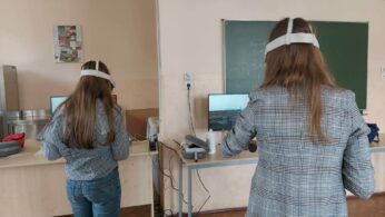 Dwie osoby z długimi włosami, ubrane w szare kurtki w kratę i dżinsy, stoją przy biurkach w klasie. Oboje używają zestawów słuchawkowych i kontrolerów wirtualnej rzeczywistości. W tle widoczna jest tablica i ekrany komputerów.