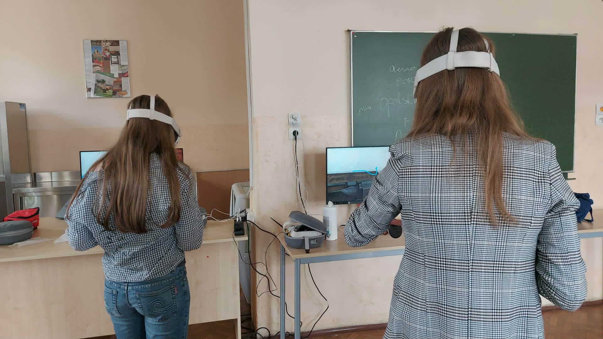 Dwie osoby z długimi włosami, ubrane w szare kurtki w kratę i dżinsy, stoją przy biurkach w klasie. Oboje używają zestawów słuchawkowych i kontrolerów wirtualnej rzeczywistości. W tle widoczna jest tablica i ekrany komputerów.