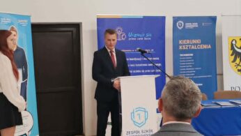 Mężczyzna w garniturze stoi na podium i wygłasza przemówienie w pomieszczeniu z drewnianą podłogą. Ludzie siedzą naprzeciwko niego. Za nim na białej ścianie wiszą banery i zegar. Powyżej widać oprawiony emblemat i nowoczesne światła. Kobieta stoi po lewej stronie obok plakatu.