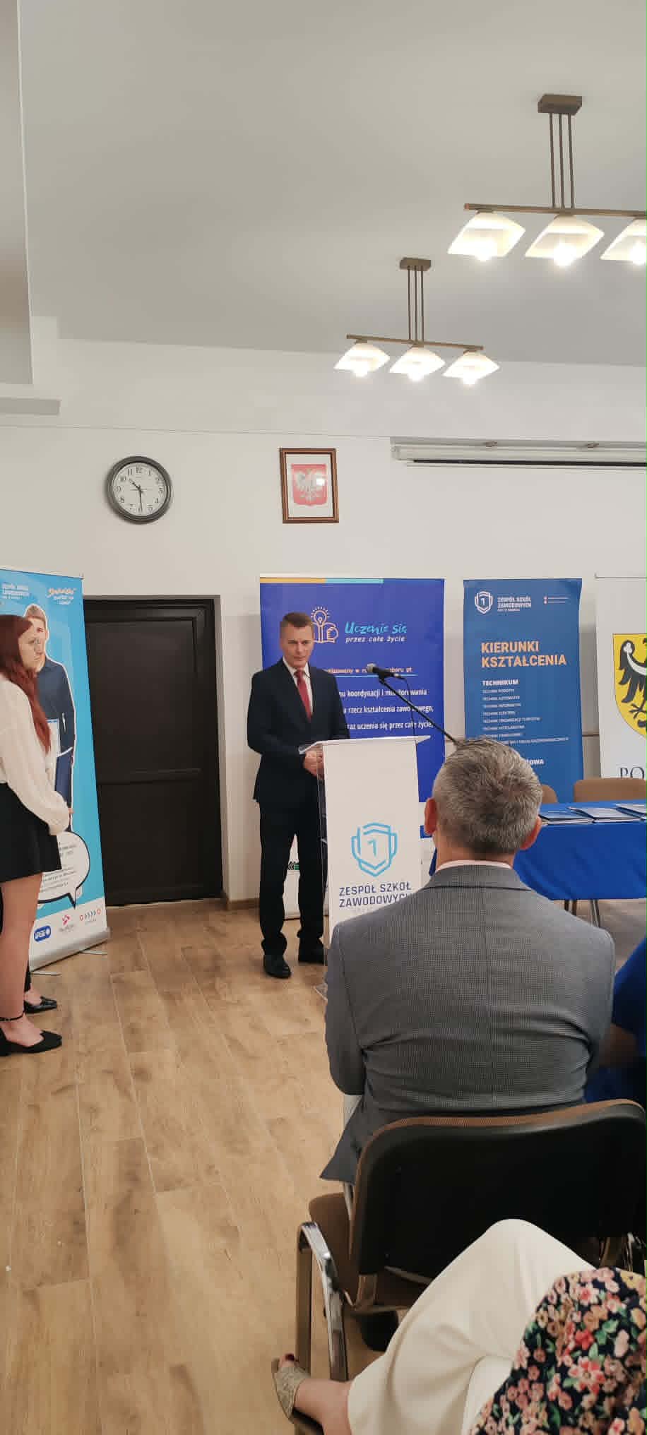 Mężczyzna w garniturze stoi na podium i wygłasza przemówienie w pomieszczeniu z drewnianą podłogą. Ludzie siedzą naprzeciwko niego. Za nim na białej ścianie wiszą banery i zegar. Powyżej widać oprawiony emblemat i nowoczesne światła. Kobieta stoi po lewej stronie obok plakatu.