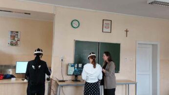 Szerokie ujęcie sali lekcyjnej, w której dwie osoby w goglach do rzeczywistości wirtualnej stoją przed monitorami pod nadzorem instruktorki. Nad zieloną tablicą wisi zegar oraz godło, a po prawej stronie widoczny jest krzyż na ścianie. Stanowiska pracy są wyposażone w komputery i nowoczesny sprzęt multimedialny.