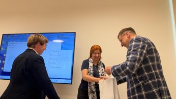 Trzy osoby znajdują się w jasnym pomieszczeniu biurowym lub konferencyjnym. Mężczyzna w kraciastej marynarce po prawej stronie przekazuje białą papierową torbę z niebieskim napisem „OPOLSKIE” kobiecie o rudych włosach, która uśmiecha się i wyciąga ręce. Po lewej stronie stoi tyłem kolejna kobieta w ciemnej marynarce, przyglądająca się tej sytuacji. W tle na ścianie wisi duży monitor wyświetlający niebieski ekran systemu operacyjnego z otwartym oknem ustawień projekcji w języku portugalskim.