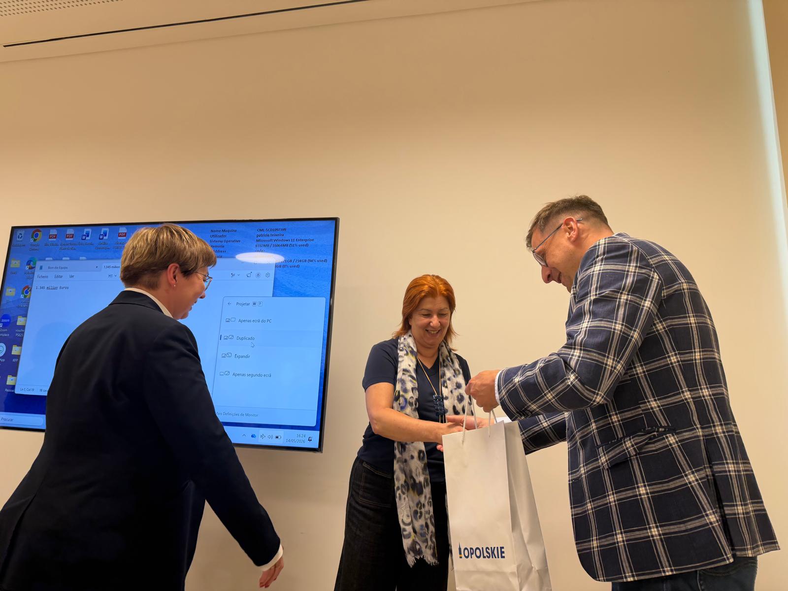 Trzy osoby znajdują się w jasnym pomieszczeniu biurowym lub konferencyjnym. Mężczyzna w kraciastej marynarce po prawej stronie przekazuje białą papierową torbę z niebieskim napisem „OPOLSKIE” kobiecie o rudych włosach, która uśmiecha się i wyciąga ręce. Po lewej stronie stoi tyłem kolejna kobieta w ciemnej marynarce, przyglądająca się tej sytuacji. W tle na ścianie wisi duży monitor wyświetlający niebieski ekran systemu operacyjnego z otwartym oknem ustawień projekcji w języku portugalskim.