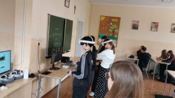 Trzech uczniów stoi przed tablicą, nosząc zestawy słuchawkowe wirtualnej rzeczywistości i stojąc przed monitorami komputerowymi na biurkach. Inni uczniowie siedzą przy biurkach w tle, pisząc lub dyskutując w grupach. Na ścianach klasy wiszą plakaty i dekoracje.