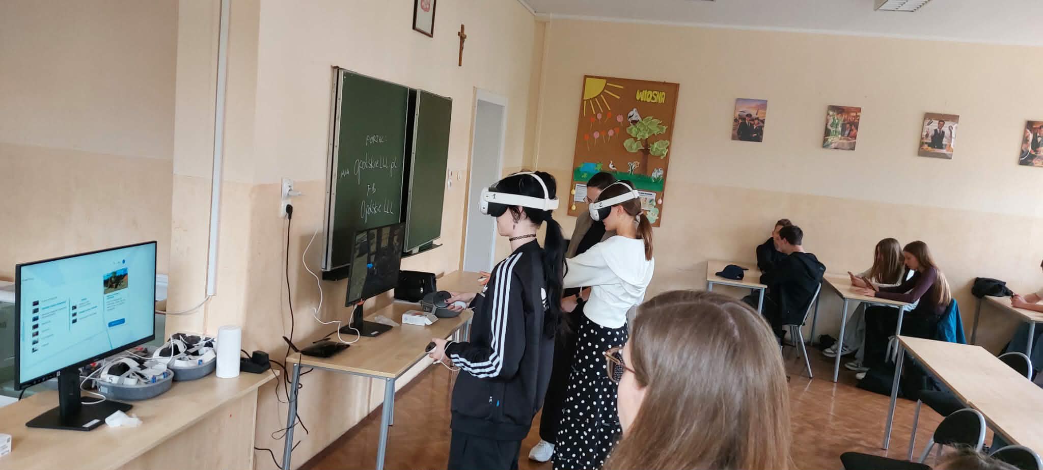Trzech uczniów stoi przed tablicą, nosząc zestawy słuchawkowe wirtualnej rzeczywistości i stojąc przed monitorami komputerowymi na biurkach. Inni uczniowie siedzą przy biurkach w tle, pisząc lub dyskutując w grupach. Na ścianach klasy wiszą plakaty i dekoracje.