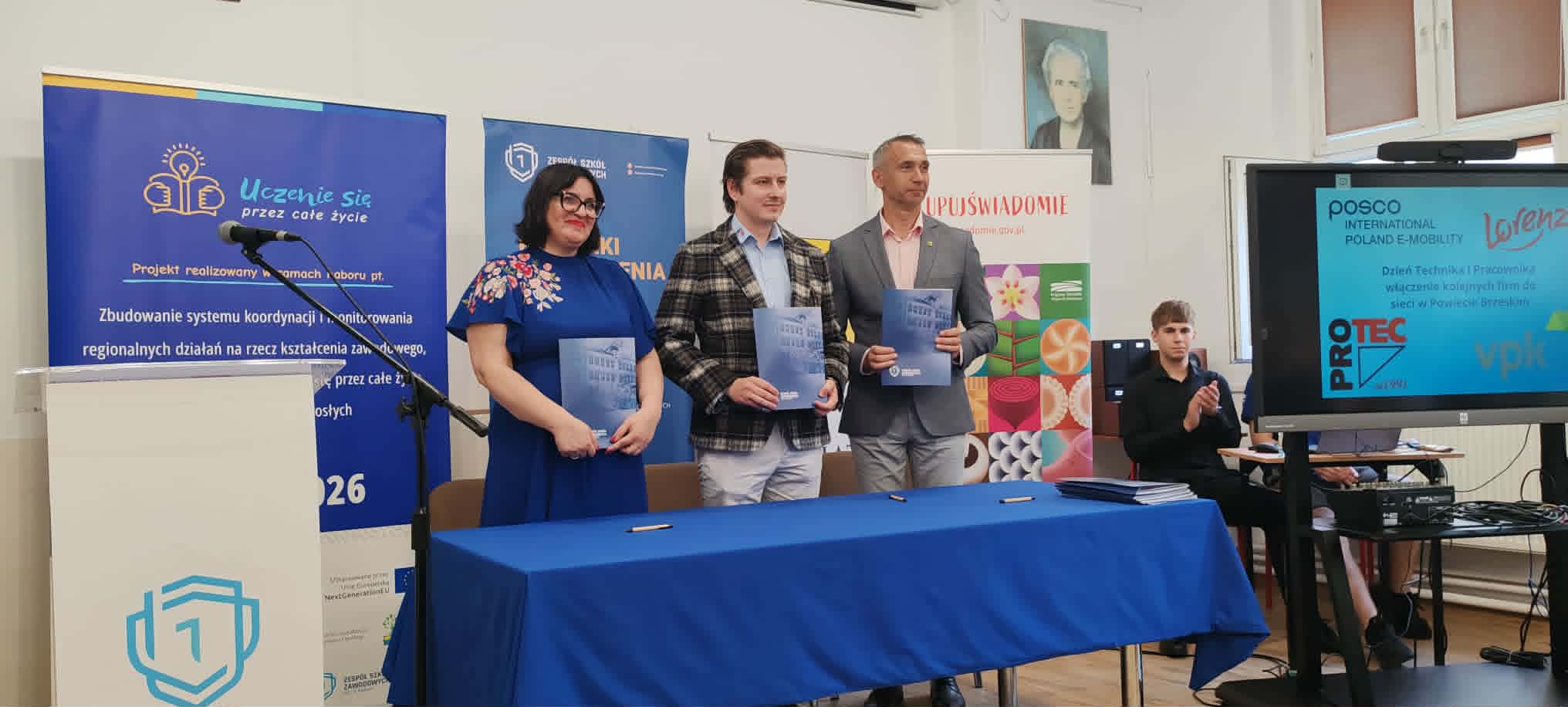 Cztery osoby stoją obok siebie trzymając niebieskie teczki, uśmiechając się, podczas gdy siedząca osoba pracuje przy laptopie. Za nimi znajdują się banery i ekran telewizora wyświetlający logo. Na ścianie wisi portret. Otoczenie wydaje się formalne, prawdopodobnie ceremonia wręczenia nagród lub certyfikatów.