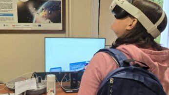 Osoba nosząca zestaw VR i niebieski plecak stoi w klasie, naprzeciwko biurka z monitorem, elektroniką i plakatami informacyjnymi o kosmosie i technologii na ścianie. Osoba wydaje się być zaangażowana w aktywność w wirtualnej rzeczywistości.