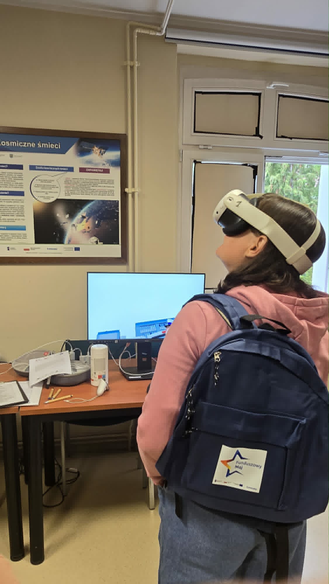 Osoba nosząca zestaw VR i niebieski plecak stoi w klasie, naprzeciwko biurka z monitorem, elektroniką i plakatami informacyjnymi o kosmosie i technologii na ścianie. Osoba wydaje się być zaangażowana w aktywność w wirtualnej rzeczywistości.