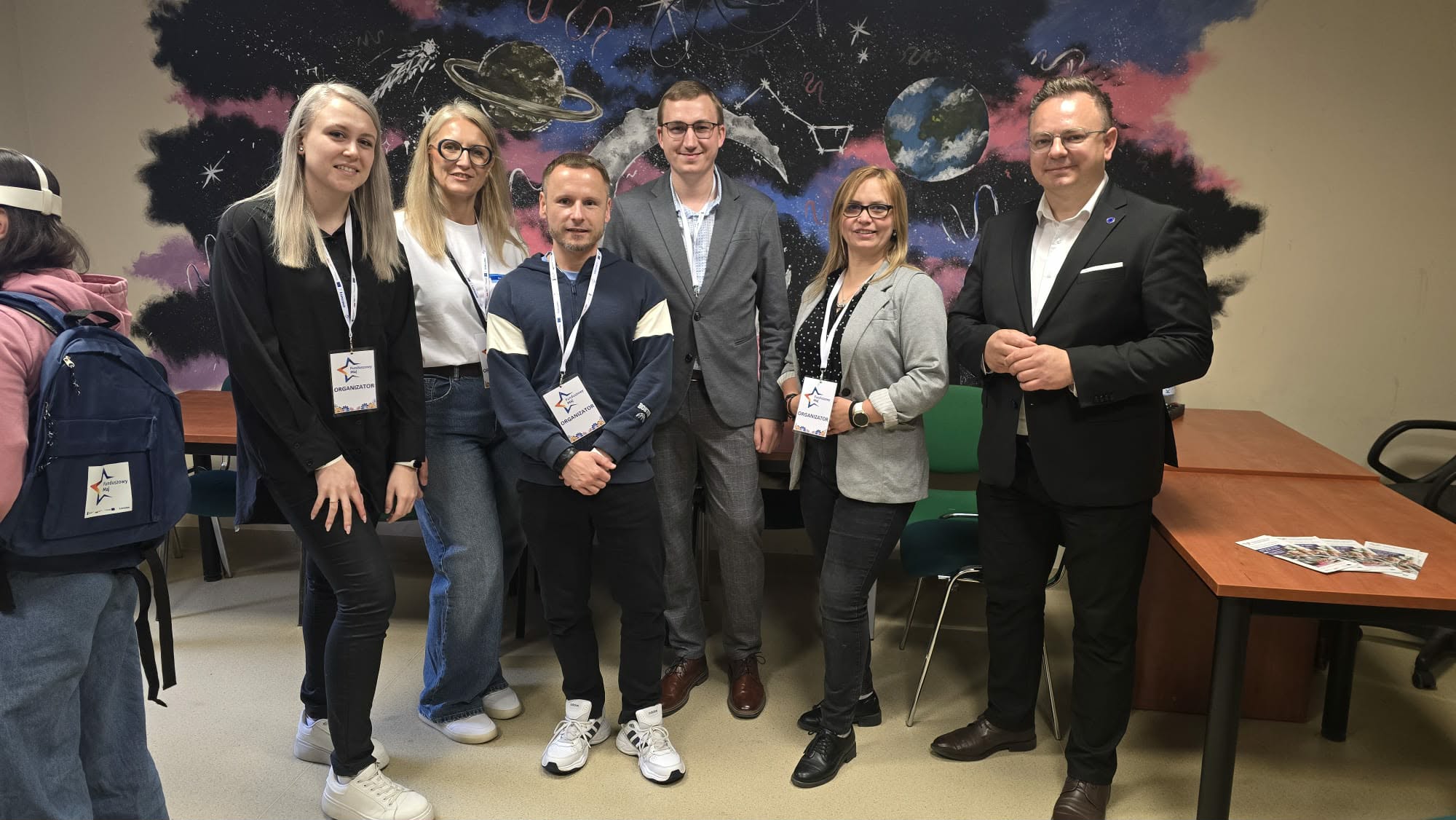 Sześć dorosłych osób pozuje w pomieszczeniu przed kolorowym muralem o tematyce kosmicznej z planetami i gwiazdami. Noszą plakietki konferencyjne i eleganckie stroje. Niektórzy stoją, inni się uśmiechają. Widoczne są stoły, krzesła i broszury. Jedna osoba z plecakiem jest częściowo poza kadrem.