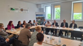 Grupa ludzi siedzi wokół dużego prostokątnego stołu w jasnej sali konferencyjnej. Większość z nich jest zaangażowana w dyskusję, a na stole leżą notatniki, kubki i butelki z wodą. Okna wpuszczają światło dzienne, a na białych ścianach wiszą banery i tablice.