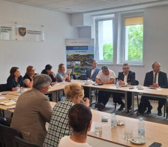 Grupa ludzi siedzi wokół dużego prostokątnego stołu w jasnej sali konferencyjnej. Większość z nich jest zaangażowana w dyskusję, a na stole leżą notatniki, kubki i butelki z wodą. Okna wpuszczają światło dzienne, a na białych ścianach wiszą banery i tablice.