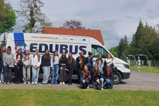 Grupa około 30 młodych ludzi stoi i klęczy obok białej furgonetki z napisem EDUBUS na utwardzonym terenie w pobliżu trawy i drzew. Niebo jest zachmurzone, a w tle widać budynek z czerwonym dachem i bramkę do piłki nożnej.