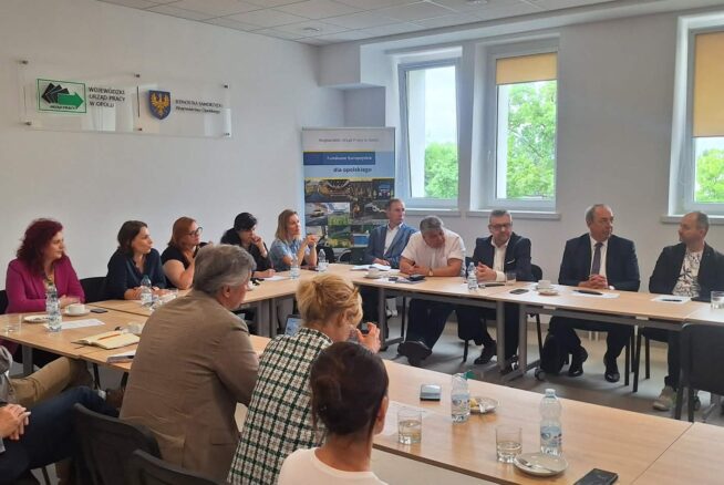 Grupa ludzi siedzi wokół dużego prostokątnego stołu w jasnej sali konferencyjnej. Większość z nich jest zaangażowana w dyskusję, a na stole leżą notatniki, kubki i butelki z wodą. Okna wpuszczają światło dzienne, a na białych ścianach wiszą banery i tablice.