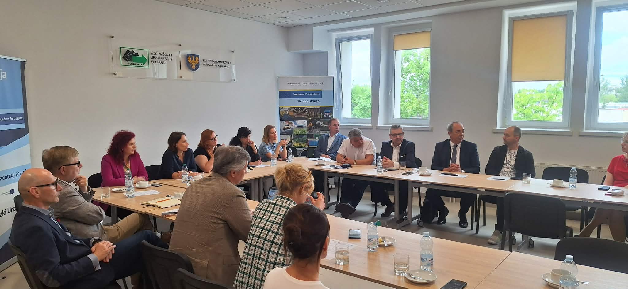 Grupa ludzi siedzi wokół dużego prostokątnego stołu w jasnej sali konferencyjnej. Większość z nich jest zaangażowana w dyskusję, a na stole leżą notatniki, kubki i butelki z wodą. Okna wpuszczają światło dzienne, a na białych ścianach wiszą banery i tablice.