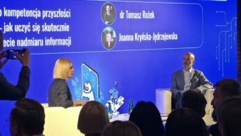 Kobieta i mężczyzna siedzą w białych fotelach na scenie podczas panelu dyskusyjnego. W tle widoczny jest duży, niebieski ekran z napisami dotyczącymi kompetencji przyszłości oraz nazwiskami prelegentów: dr Tomasz Rożek i Joanna Kryńska-Jędrzejewska. Na pierwszym planie widać sylwetki zgromadzonej publiczności tyłem do obiektywu. Po lewej stronie ktoś unosi smartfon, aby zrobić zdjęcie.