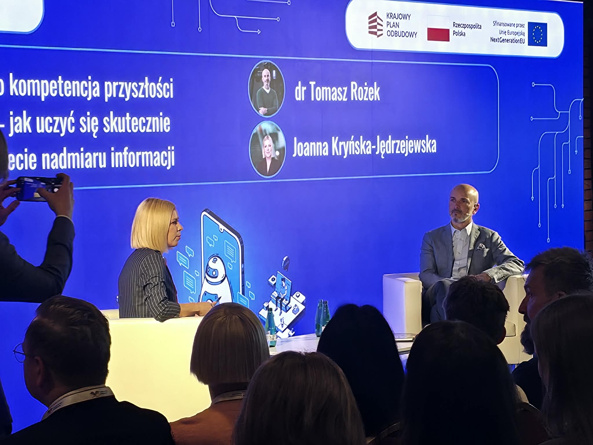 Kobieta i mężczyzna siedzą w białych fotelach na scenie podczas panelu dyskusyjnego. W tle widoczny jest duży, niebieski ekran z napisami dotyczącymi kompetencji przyszłości oraz nazwiskami prelegentów: dr Tomasz Rożek i Joanna Kryńska-Jędrzejewska. Na pierwszym planie widać sylwetki zgromadzonej publiczności tyłem do obiektywu. Po lewej stronie ktoś unosi smartfon, aby zrobić zdjęcie.