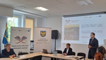 Uczestnicy spotkania w sali konferencyjnej podczas prezentacji, przy stole widoczne materiały informacyjne i ekran z wyświetlaną treścią.