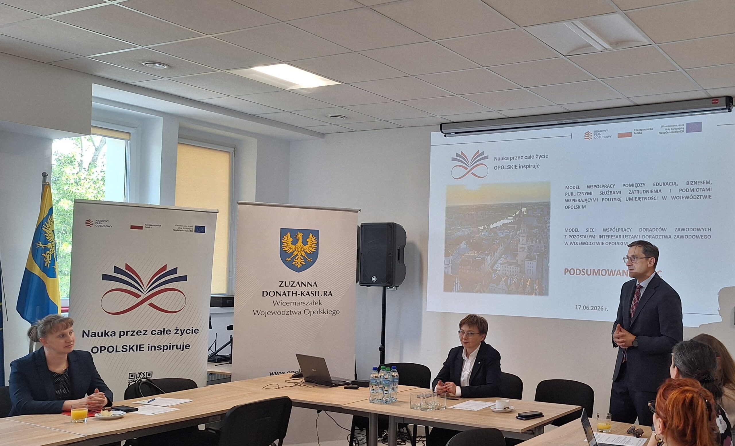 Uczestnicy spotkania w sali konferencyjnej podczas prezentacji, przy stole widoczne materiały informacyjne i ekran z wyświetlaną treścią.
