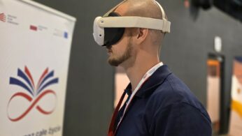 Mężczyzna z profilu, z bliska, mający na głowie białe gogle do wirtualnej rzeczywistości (VR). Ma na sobie ciemnoniebieską koszulkę polo oraz identyfikator zawieszony na szyi. W tle po lewej stronie widoczny jest fragment białego rollup-u reklamowego z napisem „...uka przez całe życie. OPOLSKIE inspiruje”. Tło za mężczyzną jest ciemnoszare i lekko rozmyte.