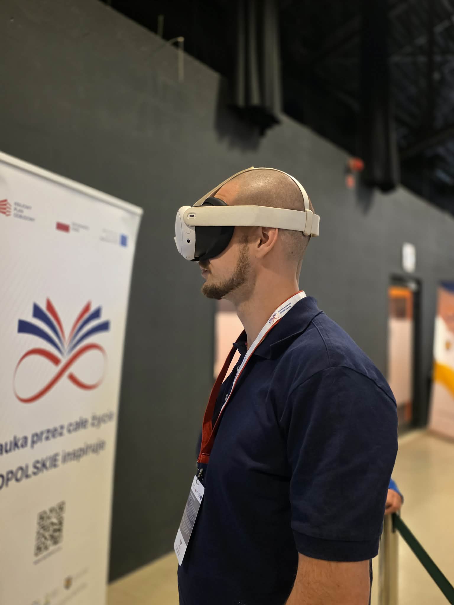 Mężczyzna z profilu, z bliska, mający na głowie białe gogle do wirtualnej rzeczywistości (VR). Ma na sobie ciemnoniebieską koszulkę polo oraz identyfikator zawieszony na szyi. W tle po lewej stronie widoczny jest fragment białego rollup-u reklamowego z napisem „...uka przez całe życie. OPOLSKIE inspiruje”. Tło za mężczyzną jest ciemnoszare i lekko rozmyte.