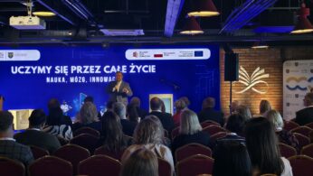 Mężczyzna w ciemnym garniturze przemawia zza mównicy podczas oficjalnego wydarzenia. W tle widoczny jest duży, jaskrawoniebieski ekran z białym napisem zawierającym słowa „CAŁE ŻYCIE” oraz logo Krajowego Planu Odbudowy i flagę Polski. Prelegent gestykuluje prawą ręką, mówiąc do mikrofonu zamontowanego na statywie. Na pierwszym planie widać zarys głowy i włosów jednej z osób siedzących na widowni.