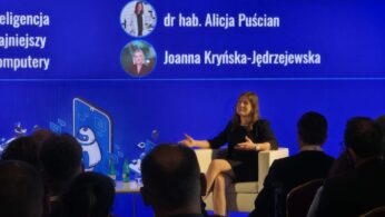 Szerokie ujęcie sali konferencyjnej wypełnionej publicznością siedzącą na krzesłach z bordową tapicerką, tyłem do obiektywu. Na scenie, przed wielkim niebieskim ekranem z napisem „UCZYMY SIĘ PRZEZ CAŁE ŻYCIE. NAUKA, MÓZG, INNOWACJE”, stoi przemawiający mężczyzna w garniturze. Na ekranie widoczne są również logotypy Unii Europejskiej oraz Krajowego Planu Odbudowy. Wnętrze ma industrialny charakter z odsłoniętym sufitem, rurami i wiszącymi lampami.