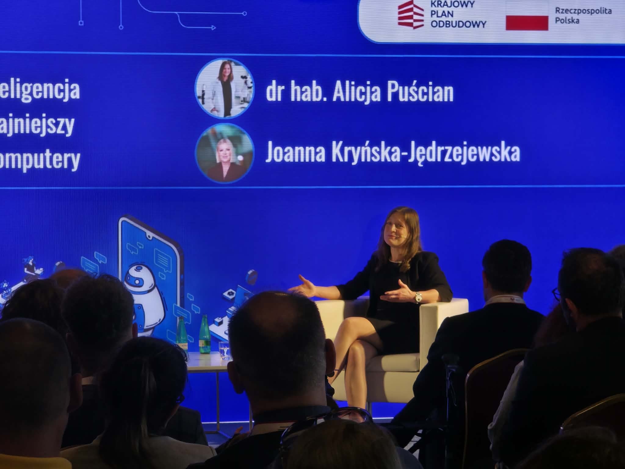 Szerokie ujęcie sali konferencyjnej wypełnionej publicznością siedzącą na krzesłach z bordową tapicerką, tyłem do obiektywu. Na scenie, przed wielkim niebieskim ekranem z napisem „UCZYMY SIĘ PRZEZ CAŁE ŻYCIE. NAUKA, MÓZG, INNOWACJE”, stoi przemawiający mężczyzna w garniturze. Na ekranie widoczne są również logotypy Unii Europejskiej oraz Krajowego Planu Odbudowy. Wnętrze ma industrialny charakter z odsłoniętym sufitem, rurami i wiszącymi lampami.