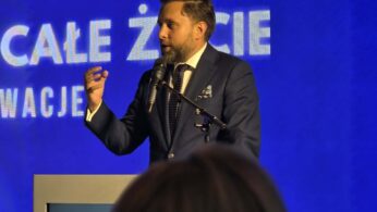 Kobieta w ciemnym stroju siedzi w białym fotelu na scenie podczas panelu dyskusyjnego i gestykuluje rękami w trakcie wypowiedzi. Przed sceną widoczne są tyły głów publiczności uczestniczącej w spotkaniu. W tle znajduje się duży niebieski ekran multimedialny wyświetlający m.in. grafikę z robotem oraz napisy „dr hab. Alicja Puścian” i „Joanna Kryńska-Jędrzejewska” wraz z ich okrągłymi zdjęciami profilowymi. W prawym górnym rogu ekranu widnieje logo Krajowego Planu Odbudowy.