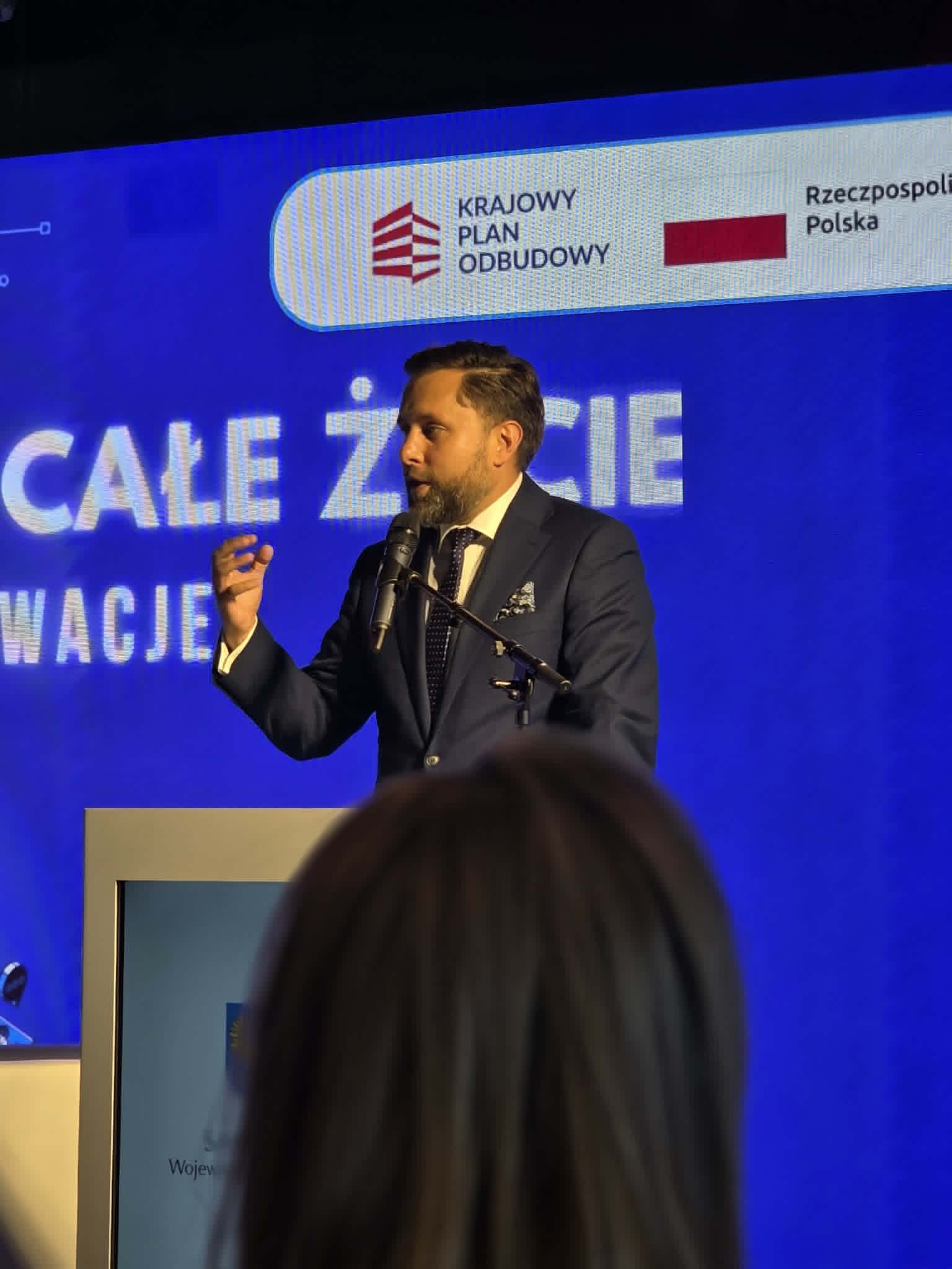 Kobieta w ciemnym stroju siedzi w białym fotelu na scenie podczas panelu dyskusyjnego i gestykuluje rękami w trakcie wypowiedzi. Przed sceną widoczne są tyły głów publiczności uczestniczącej w spotkaniu. W tle znajduje się duży niebieski ekran multimedialny wyświetlający m.in. grafikę z robotem oraz napisy „dr hab. Alicja Puścian” i „Joanna Kryńska-Jędrzejewska” wraz z ich okrągłymi zdjęciami profilowymi. W prawym górnym rogu ekranu widnieje logo Krajowego Planu Odbudowy.