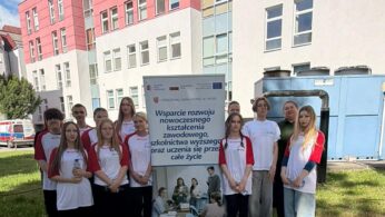 Grupa uczestników wydarzenia edukacyjnego przed budynkiem szpitala, trzymająca tablicę informacyjną