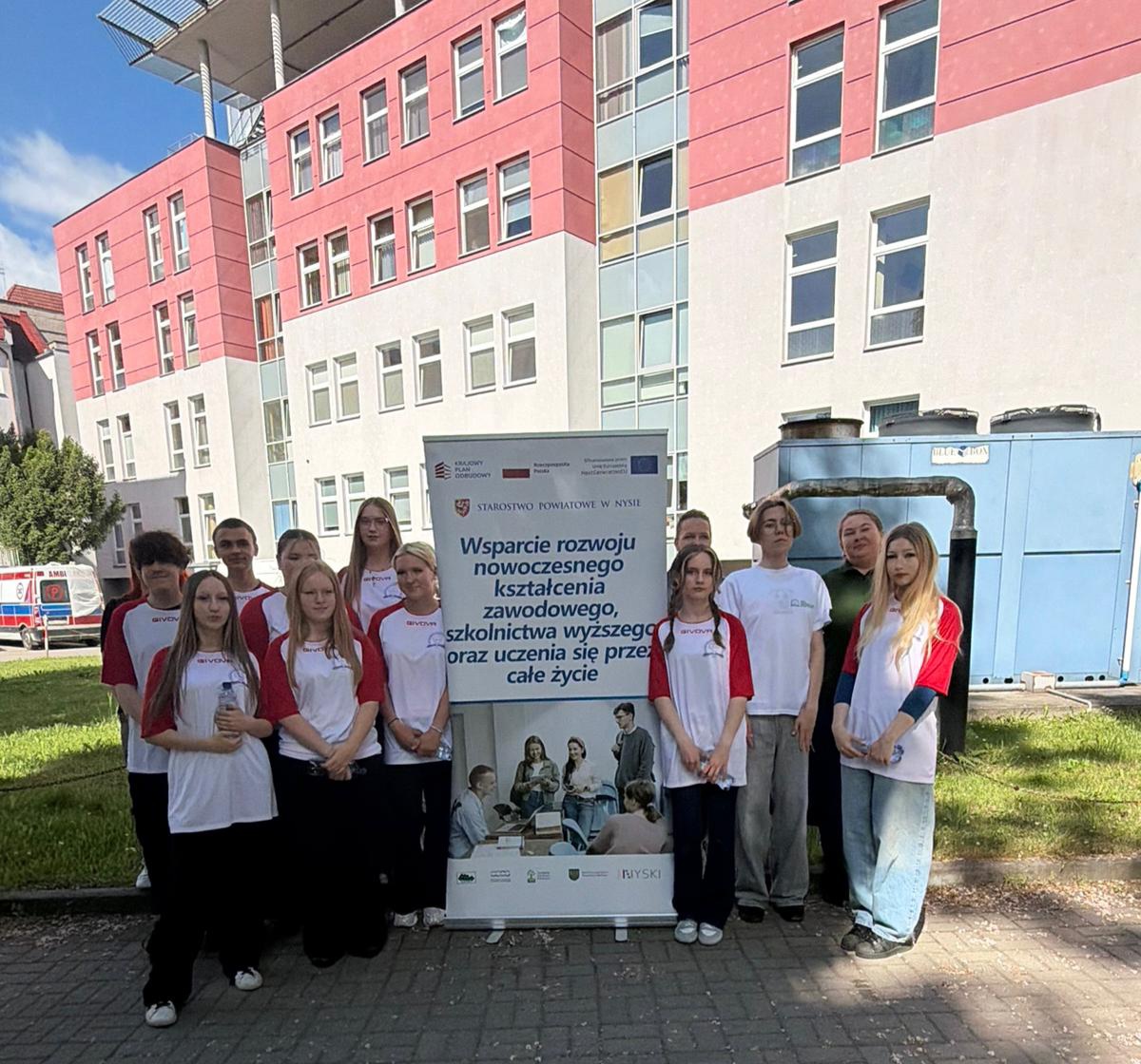 Grupa uczestników wydarzenia edukacyjnego przed budynkiem szpitala, trzymająca tablicę informacyjną