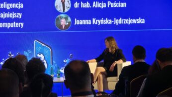 Kobieta siedzi w białym fotelu na scenie podczas konferencji, gestykulując w trakcie wypowiedzi. Za nią znajduje się wielki, niebieski ekran multimedialny prezentujący temat dotyczący sztucznej inteligencji oraz komputerów. Na ekranie wyświetlają się także nazwiska: dr hab. Alicja Puścian oraz Joanna Kryńska-Jędrzejewska. Z przodu widoczne są ciemne sylwetki słuchaczy siedzących na widowni.