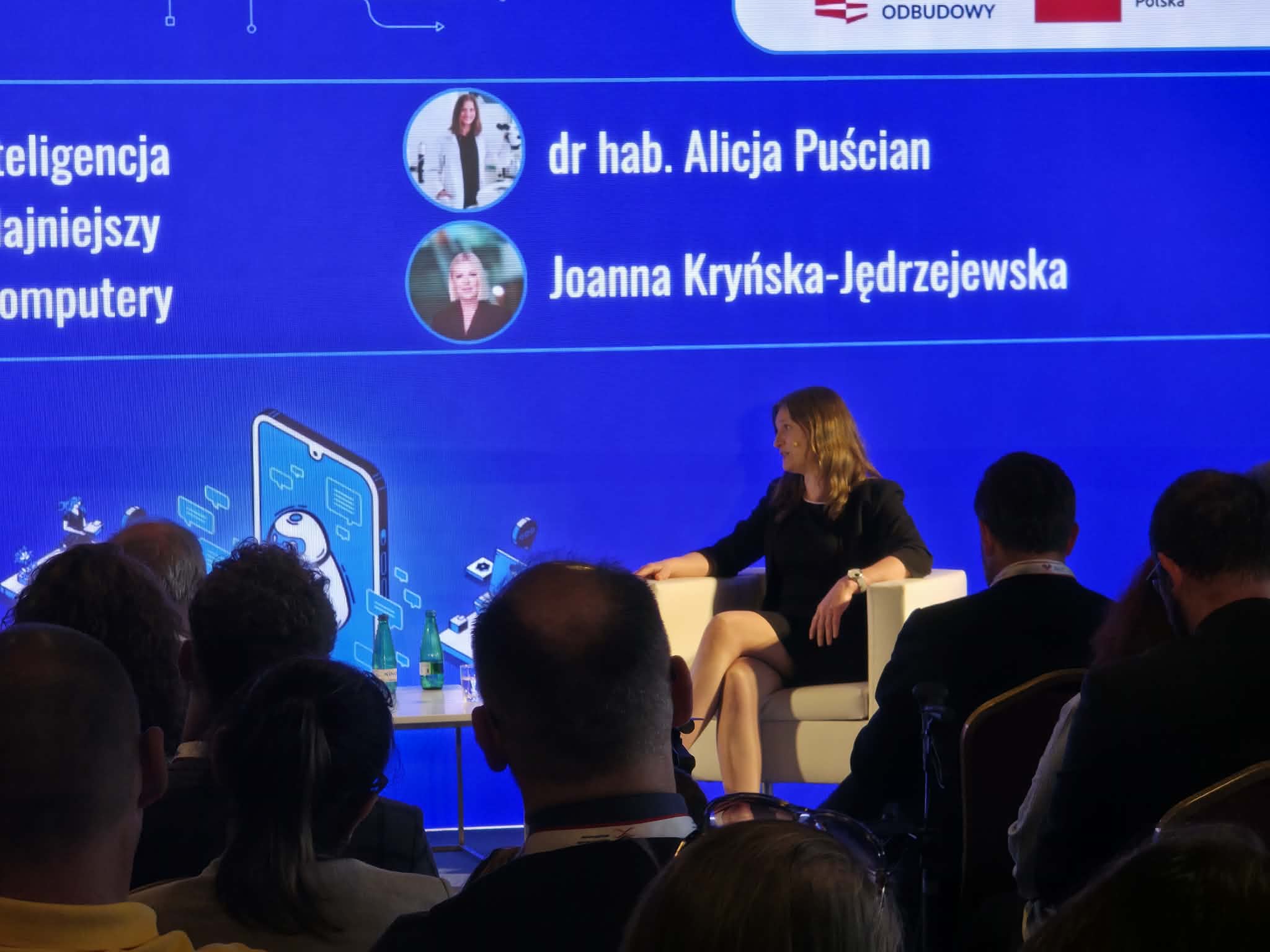 Kobieta siedzi w białym fotelu na scenie podczas konferencji, gestykulując w trakcie wypowiedzi. Za nią znajduje się wielki, niebieski ekran multimedialny prezentujący temat dotyczący sztucznej inteligencji oraz komputerów. Na ekranie wyświetlają się także nazwiska: dr hab. Alicja Puścian oraz Joanna Kryńska-Jędrzejewska. Z przodu widoczne są ciemne sylwetki słuchaczy siedzących na widowni.